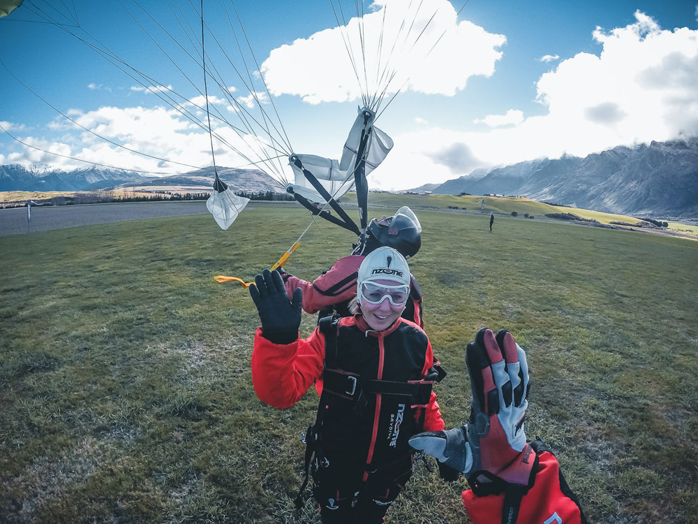 Girl wearing a skydiving jumpsuit an cap giving a high five to a gloved hand with a white parachute floating behind them right after landing in a green field surrounded by mountains.