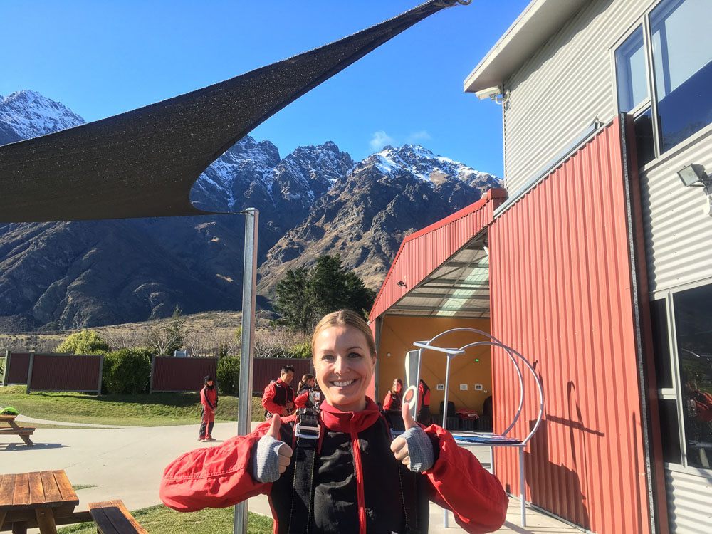 Girl wearing a skydiving jumpsuit giving two thumbs up with mountains in the background just before getting on plane to go skydiving in Queenstown.