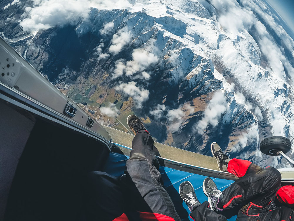 Two pairs of legs belonging to skydivers dangling over the side of an open door small aircraft with snow-capped mountains below.