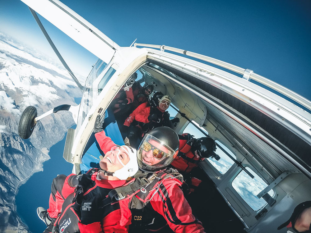 Tandem skydivers about to jump out of small plane on a clear day with snow-capped mountains below.