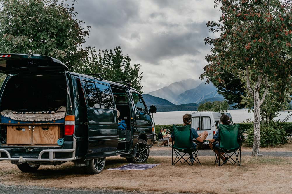 Two people sitting in camping chairs next to a camper van in front of a lake