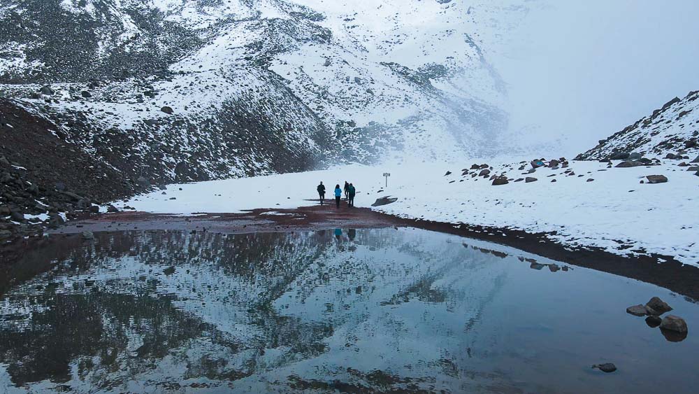 People standing on the edge of an alpine lake surrounded by snow covered mountains