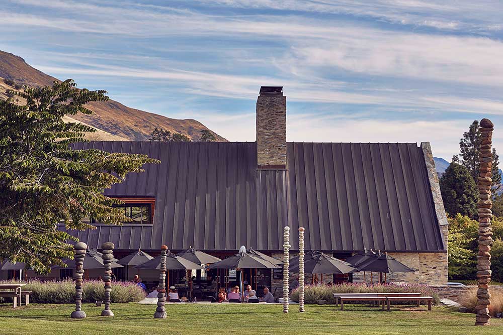 Exterior view of Amisfield Winery in Queenstown on a fine day, featuring a stone building with a brown sloped roof and people sitting at umbrella covered tables in outdoor patio.