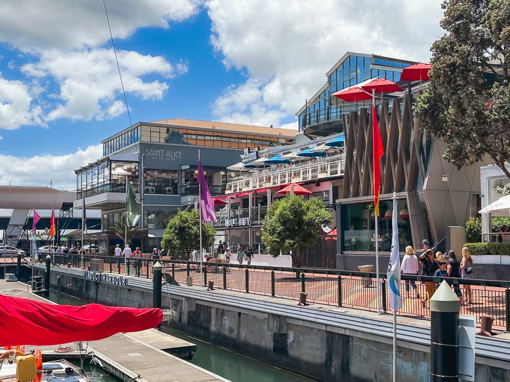 People walking along Viaduct Harbour in Auckland with bars and restaurants lining the promenade on a summer's day.