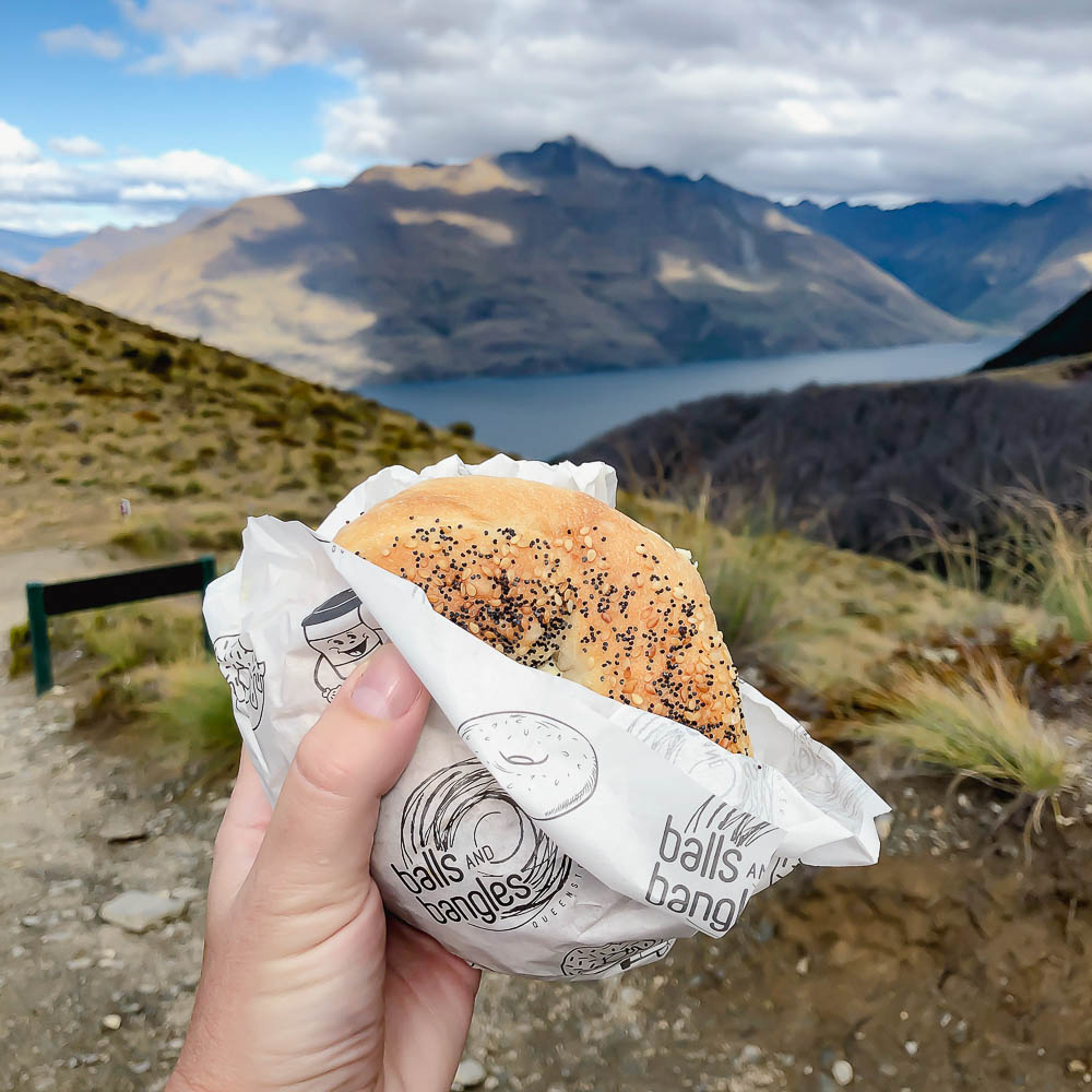 hand holding a bagel wrapped in paper with a mountain in the background