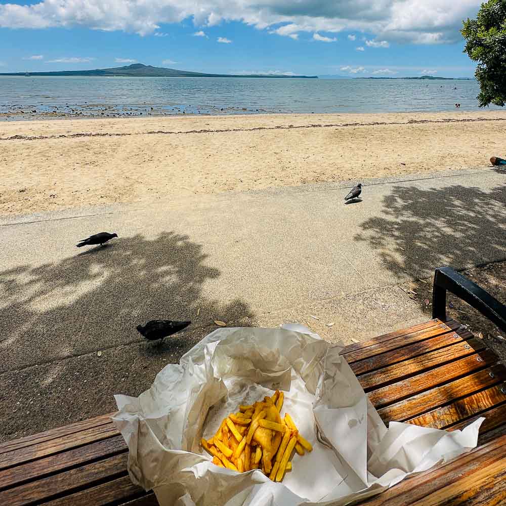 fish and chips in paper sitting on a park bench in front of the ocean on a clear day.