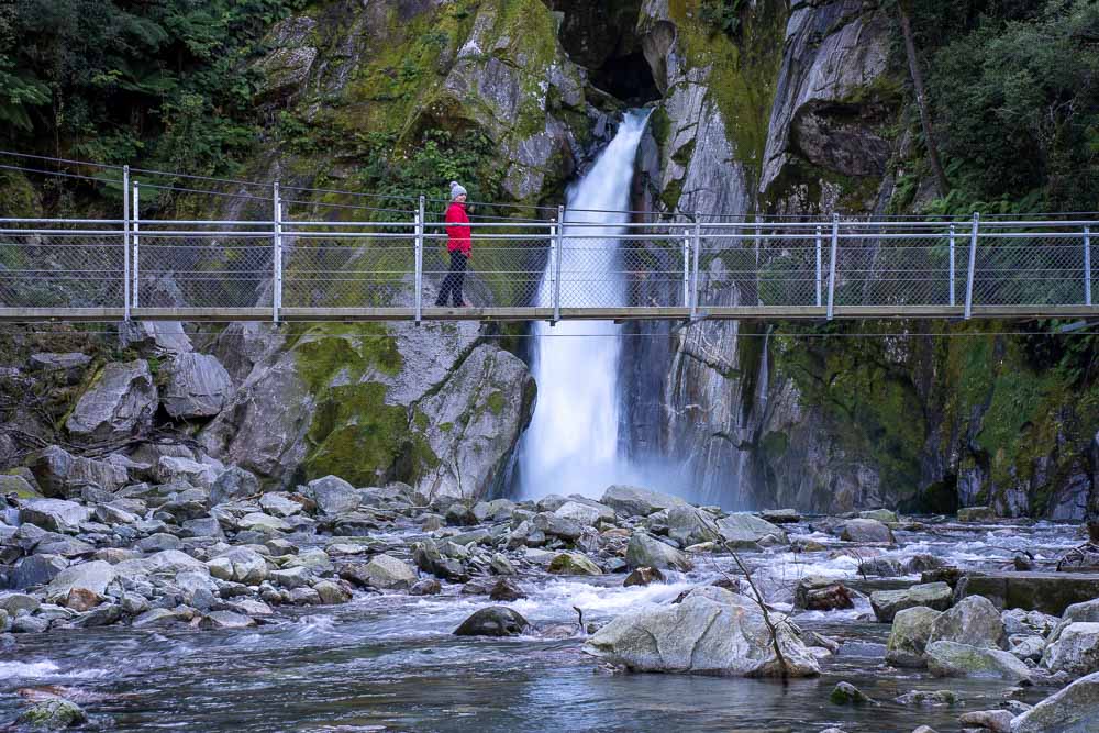 A girl in a red jacket walking across a swing bridge in front of Giant Gate Falls on the Milford Track in New Zealand.