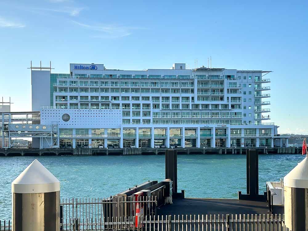 Exterior of the Hilton Hotel a white multi-story rectangular building surround by the blue harbor on three sides.