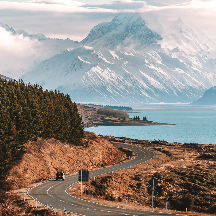 Black car driving along the winding road to Mt Cook alongside a blue lake with a snow capped mountain in the distance.