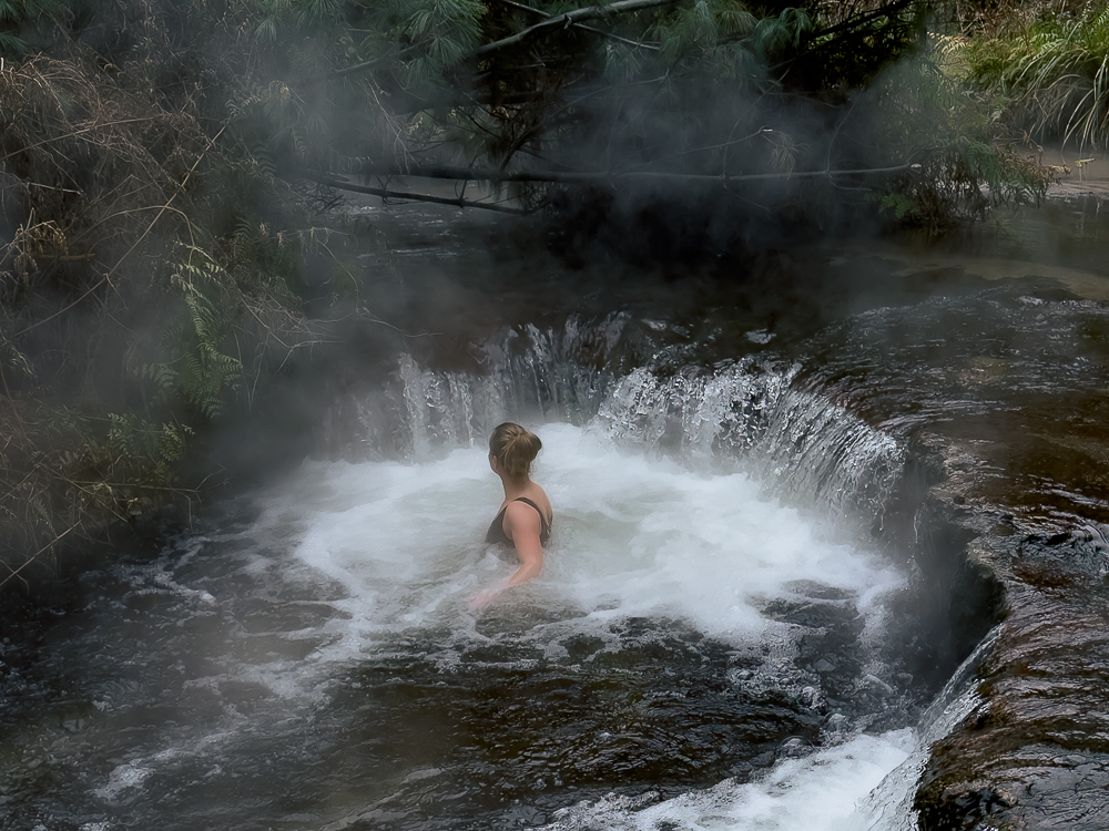 Woman soaking in a small natural hot pool in the forest called Kerosene Creek with steam rising from the water in Rotorua New Zealand.
