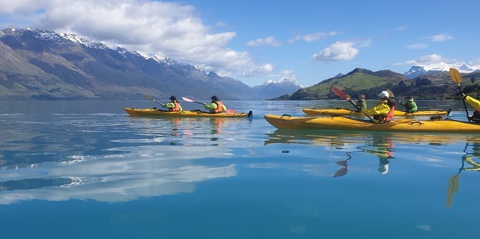 A group of people kayaking on the calm blue water of a lake surrounded by mountains in New Zealand.