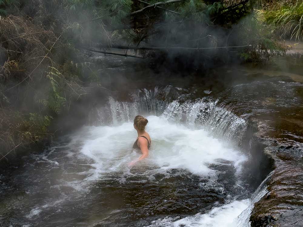 Woman soaking in a small natural hot pool in the forest called Kerosene Creek with steam rising from the water in Rotorua New Zealand.