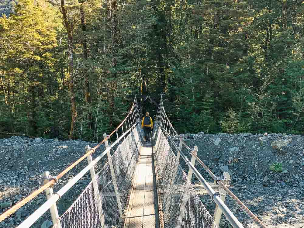 Girl in yellow shirt and black backpack walking over a swingbridge and into the forest in Glenorchy.