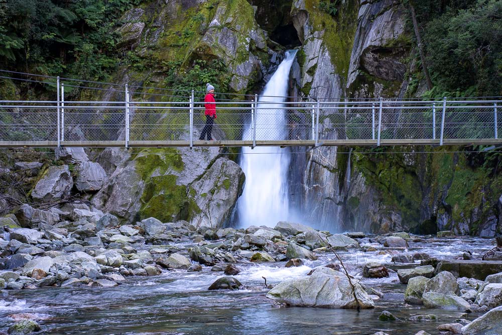 girl in red jacket walking on swing bridge in front of waterfall