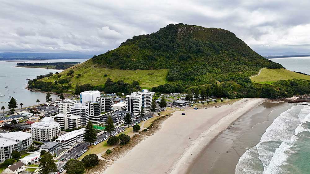 Aerial view of the long white sands of Mount Maunganui beach lined with shops and the lush green cone of Mount Maunganui volcano at the end of the beach. 