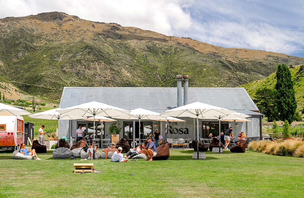 A group of people drinking wine while sitting in beanbags shaded by white umbrellas on the front lawn of Mt Rosa's tasting room with a mountain in the background on a fine summer day. 