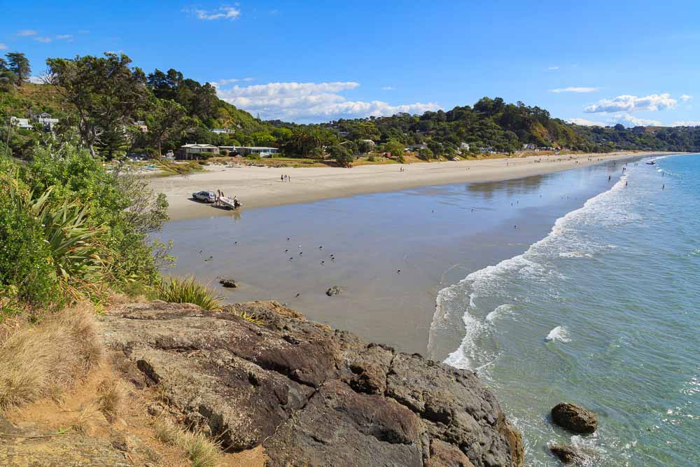A few people enjoying the white sands of Onetangi Beach on Waiheke Island as seen from the top of a rock on a clear day.