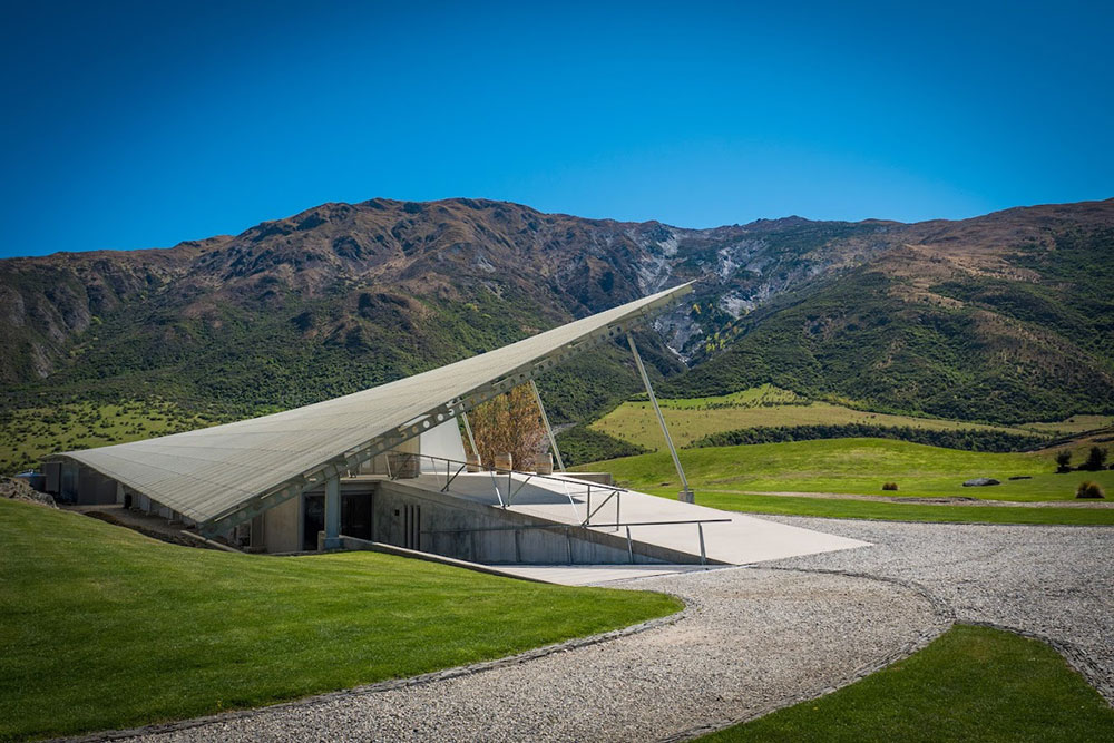 The uniquely shaped building of Peregrine Wines in Otago New Zealand, surrounded by green grass and mountains on a clear day.