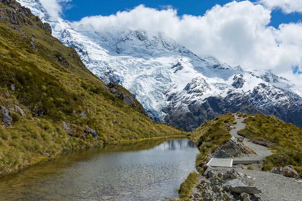 A gravel walking path along the Sealy Tarns with large snow capped mountains in the background in Mt Cook National Park.