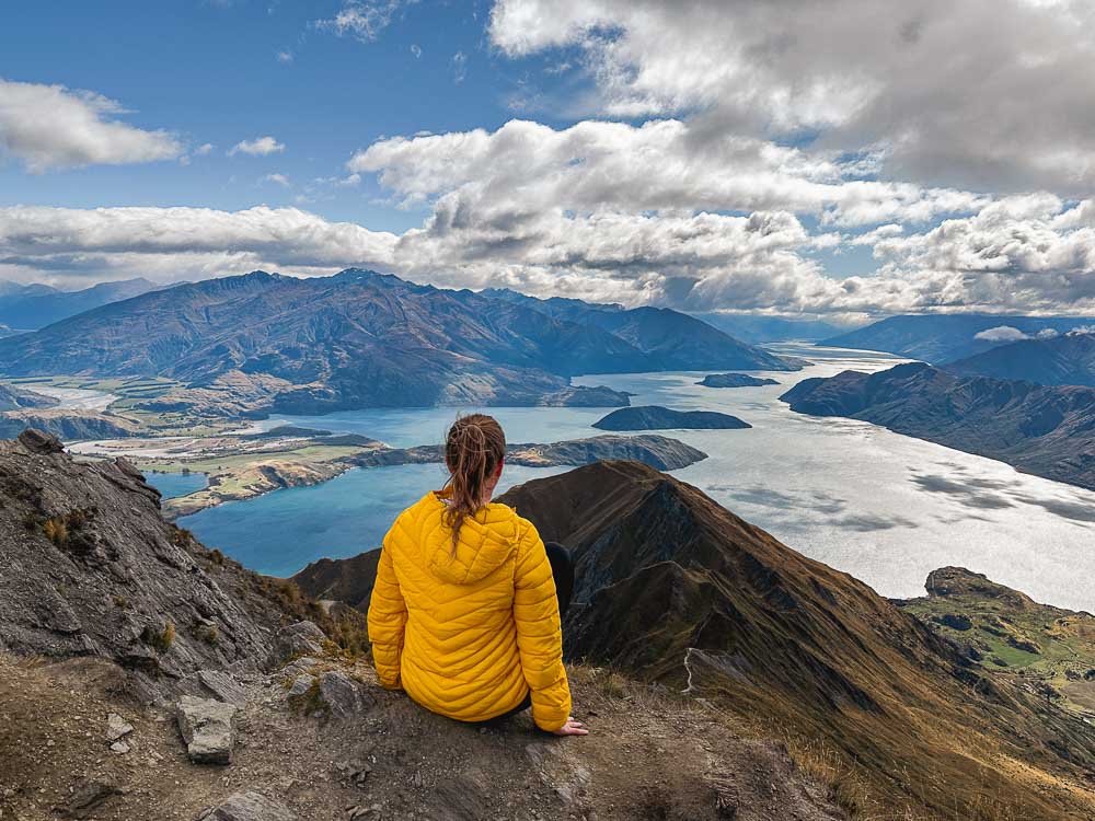 girl in yellow jacket sitting on top of a mountain with large lake in the background