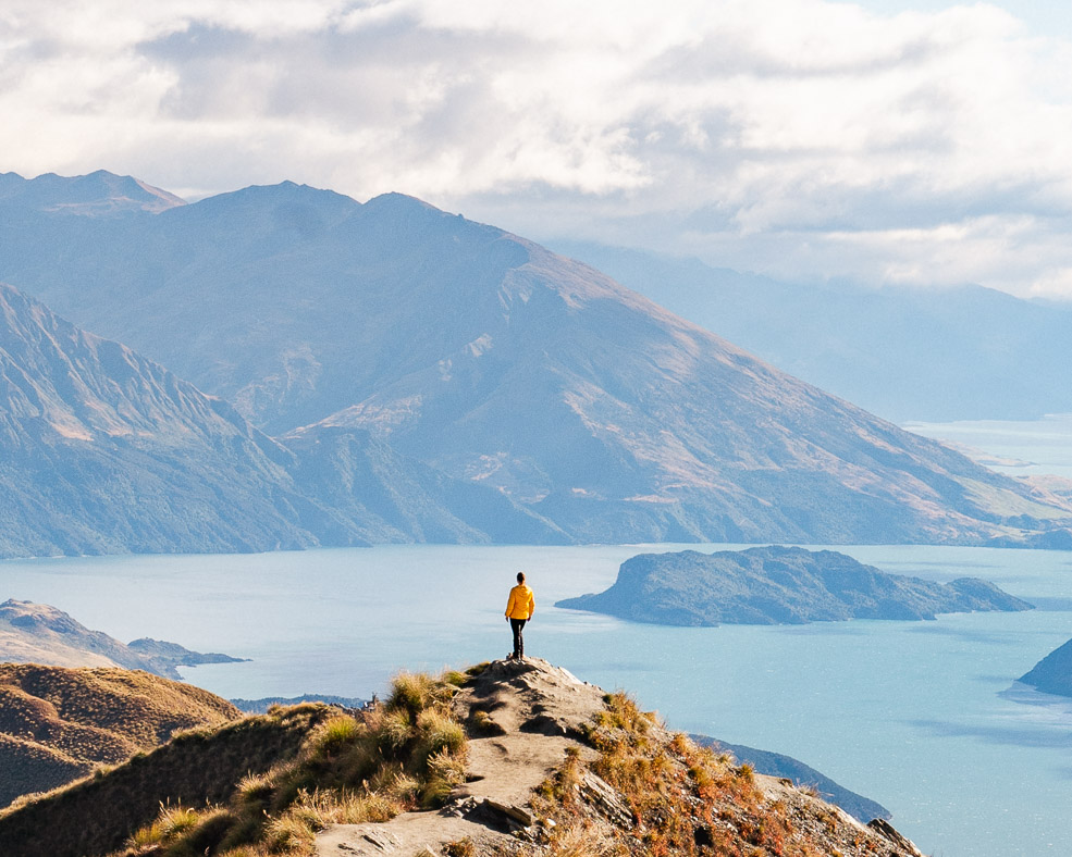 girl in yellow jacket standing on top of mountain with mountain and a lake in the background