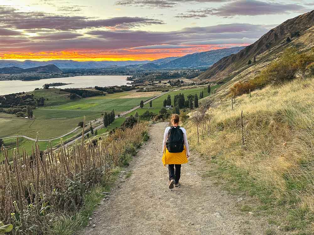 girl with black backpack walking down hill with sunrise in the background