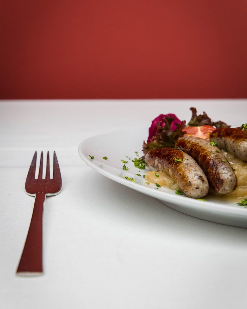 A close up of two sausages on a white plate with a silver fork to represent the small town of Tuatapere who claims to be the sausage capital of New Zealand.