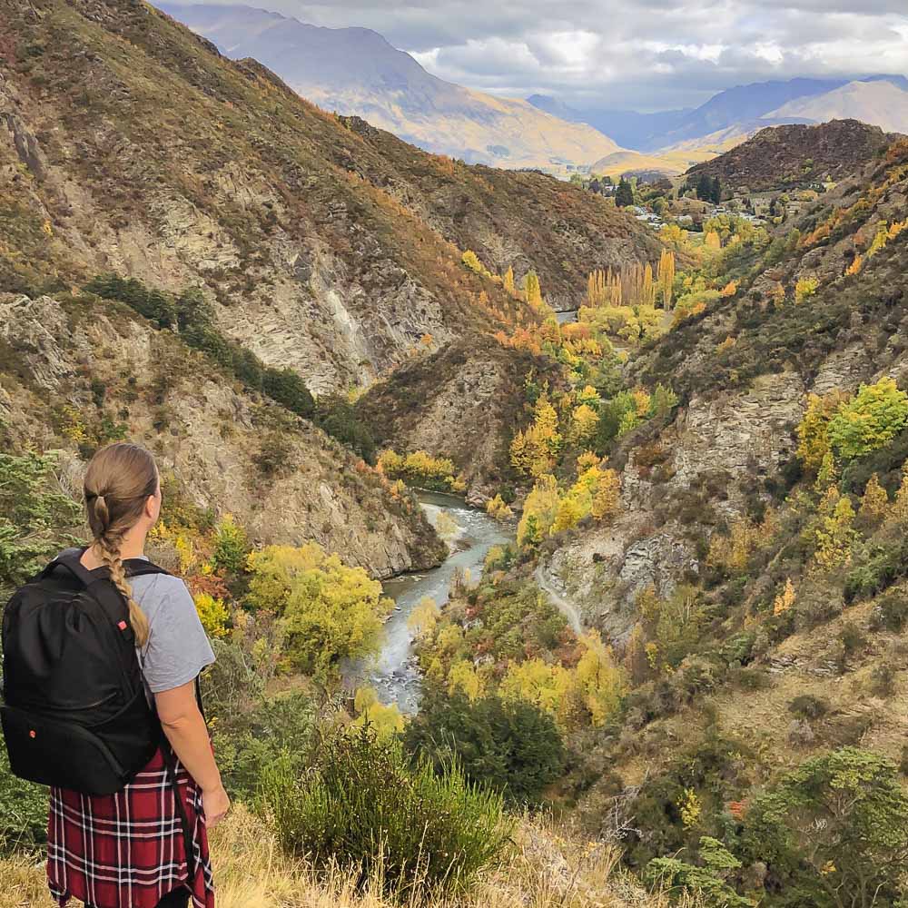 Girl wearing a black backpack standing over a valley filled with fall colors on the Sawpit Gully Track in Arrowtown New Zealand.