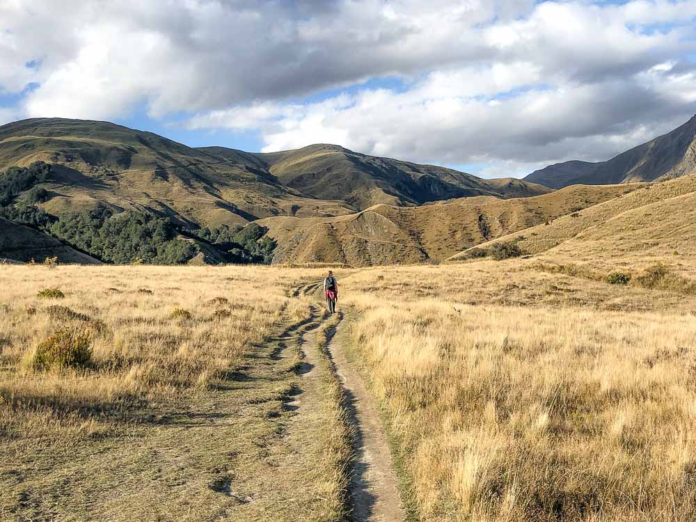 Girl walking on a path through a tussock field with rolling hills in the distance on a partly cloudy day,