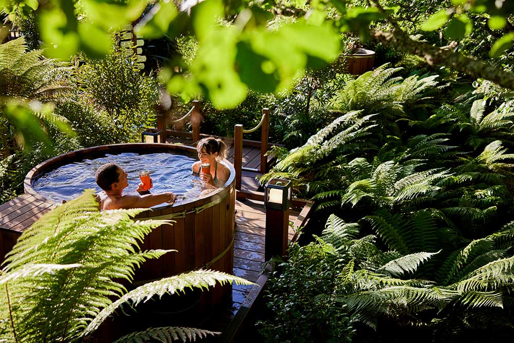 A couple relaxing in a wooden hot tub surrounded by green native plants at the Secret Spot in Rotorura, New Zealand.