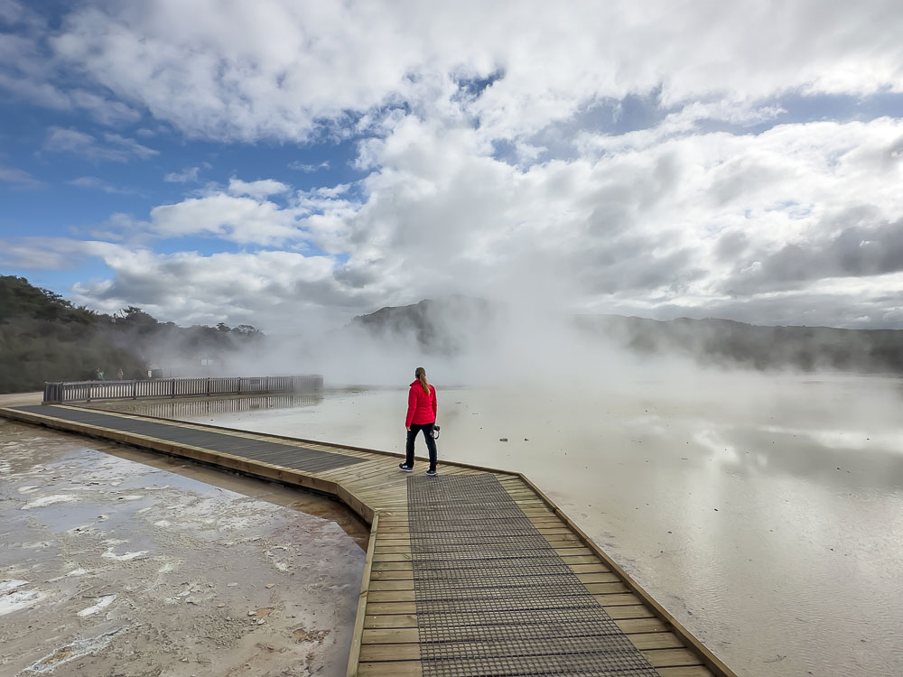 Person wearing a red jacket walking along a boardwalk through bubbling mud and steam at Wai o Tapu Thermal Wonderland, an experience worth adding to your Rotorua itinerary.