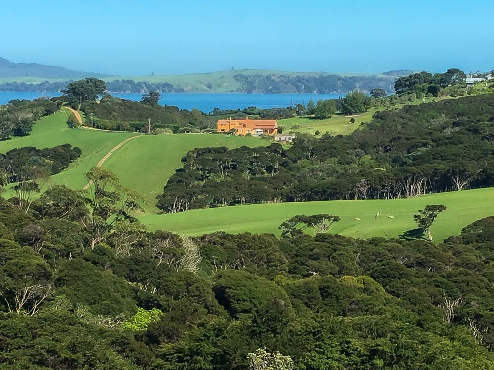 A large rustic terracotta house on Waiheke Island surrounded by green trees and grass fields with the blue harbor water behind it.