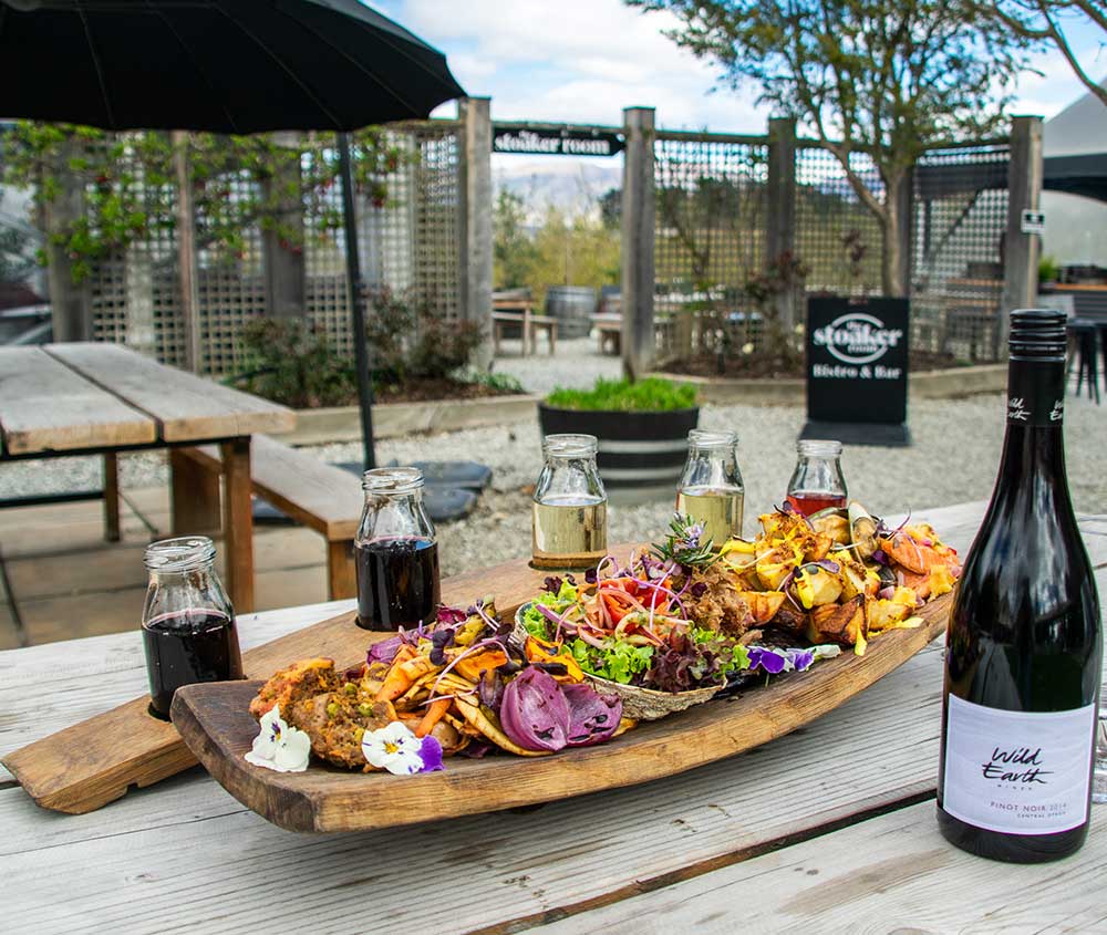 A wooden platter filled with fresh New Zealand cuisine along with a flight of Wild Earth Wines and a bottle of Wild Earth Pinot Noir.